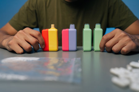 Cropped shot of person organizing vape pods, prescription pills and powder on table during nighttime preparation, close-up. Concept of illegal substance.の写真素材