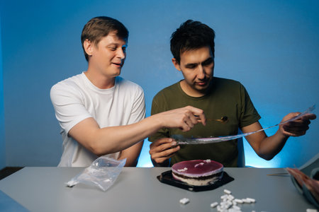 Two people mixing substances inside a room, preparing a cake with layered decoration, under blue...