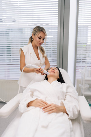 Vertical portrait of cosmetologist female performing microneedling anti-aging skincare treatment on woman face in modern beauty clinic, promoting rejuvenation. Concept of beauty, facial antiaging.の写真素材