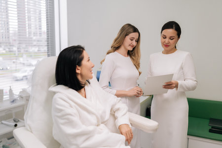 Two professional cosmetologists consulting client during beauty clinic visit, discussing treatment option, providing professional skincare and wellness service. Concept of skin care treatment.の写真素材