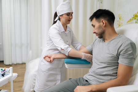 Side view of medical worker in uniform and gloves preparing equipment for drawing blood from male patient sitting in examination chair in modern clinic for health checkup.の写真素材