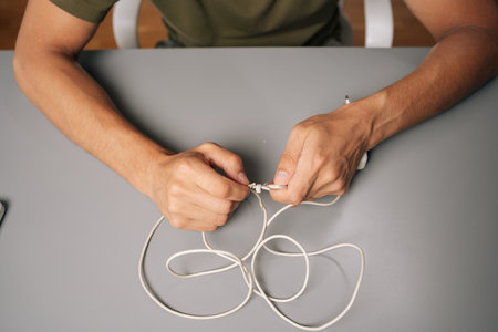 Close-up of technician hands tying knot in damaged white charger cable, attempting repair at gray desk, demonstrating resourcefulness and problem-solving skills. Concept of computer hardwareの写真素材