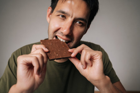 Close-up face of happy young man sitting at table biting chocolate, savoring rich flavor of dark chocolate bar, blissful enjoyment as takes satisfying and delicious bite, on isolated background.の写真素材