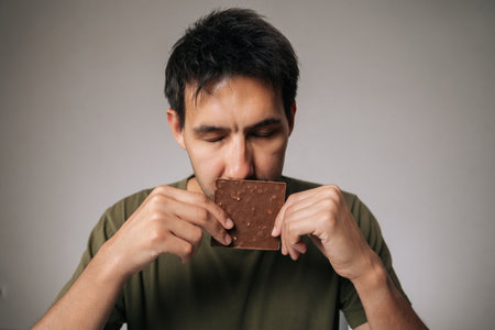 Studio portrait of enjoying young man savoring rich aroma of dark chocolate bar with closed eyes, holding it close to face in anticipation of delightful treat, sitting at table on isolated background.の写真素材
