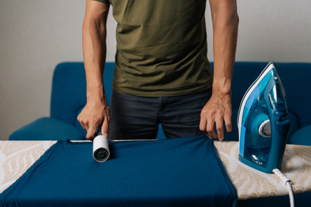 Cropped shot of man using sticky lint roller to remove dust and lint from blue t-shirt on ironing board, with steam iron nearby, preparing to iron and care for clothes at home. Concept of housekeepingの写真素材
