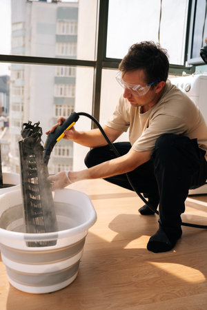 Vertical shot of professional cleaning service worker wearing safety glasses using steam cleaner to clean air conditioner filter in bucket of water, performing maintenance on HVAC system.の写真素材