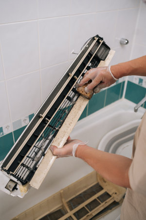 Close-up cropped shot of technician wearing gloves carefully cleaning dirty air conditioner filter with sponge and soapy water, ensuring proper hygiene and improved air quality.の写真素材