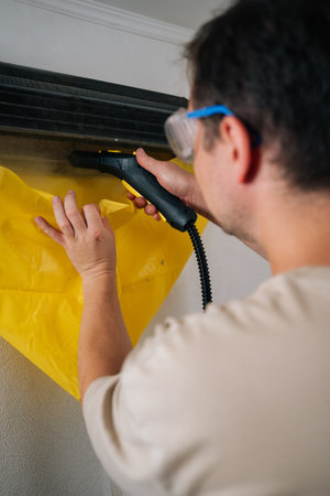 Close-up back view of technician in safety glasses cleaning air conditioner using steam cleaner while holding yellow protective sheet to collect dirt water. Concept of hygiene and cleanliness at home.の写真素材