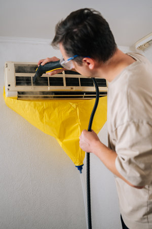 Vertical shot of professional technician in safety glasses standing on ladder and cleaning air conditioner unit using steam cleaner, with yellow plastic sheet protecting wall from dirty water.の写真素材