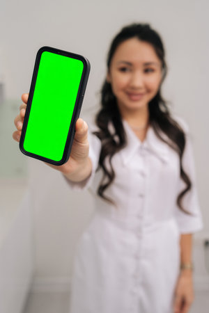 Asian cosmetologist presenting smartphone with green screen for mobile application display, offering mockups for product showcases, digital healthcare services, and beauty industry marketing.の写真素材