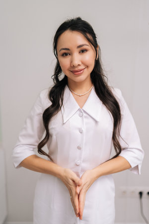 Smiling Asian woman beautician standing in white uniform, representing confident and professional cosmetologist or doctor offering beauty and aesthetic services in modern medical clinic.の写真素材