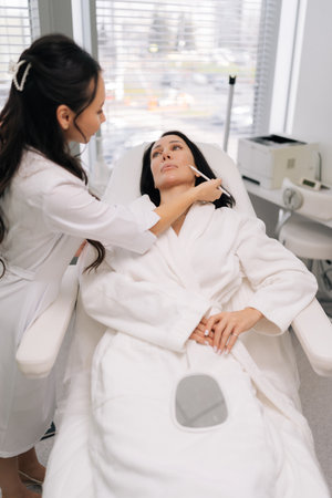 Professional beauty technician marking woman face with white pencil during pre-procedure consultation in clinic, explaining planned injection or cosmetic treatment areas. Concept of facial anti-aging.の写真素材