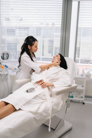 Young beauty technician marking woman face with white pencil during pre-procedure consultation in clinic, explaining planned injection or cosmetic treatment areas. Concept of facial anti-aging.の写真素材