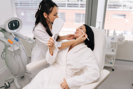 Young cosmetologist talking with female client and using white pencil to mark lines on face before cosmetic procedure, preparing for plastic surgery or beauty injections. Concept of facial anti-aging.の写真素材