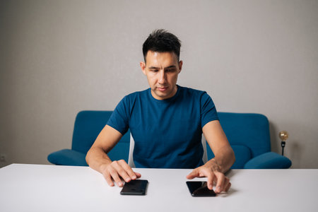 Portrait of young blogger male comparing two smartphones, making decision about which one to buy or use, sitting at white table. Concept of choosing best option.の写真素材