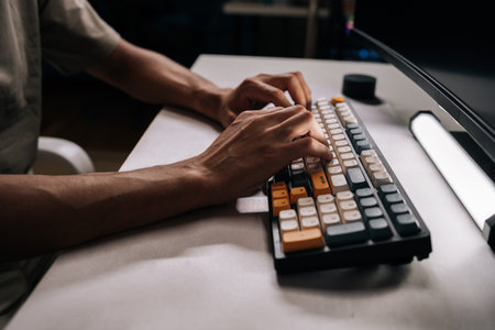 Side view of male hands typing on mechanical keyboard with colorful keycaps in dimly lit office, suggesting focused work or late-night coding session. Concept of modern technology.の写真素材
