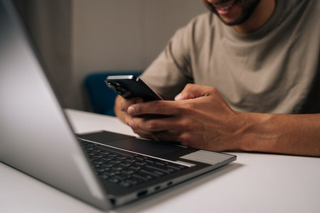 Smiling young businessman working late at night in home office, using smartphone and laptop to...