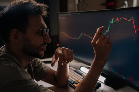 Positive young man smiling and pointing at fluctuating financial graph on desktop screen, actively managing cryptocurrency investments and tracking market trends for profitable trading decisions.の写真素材