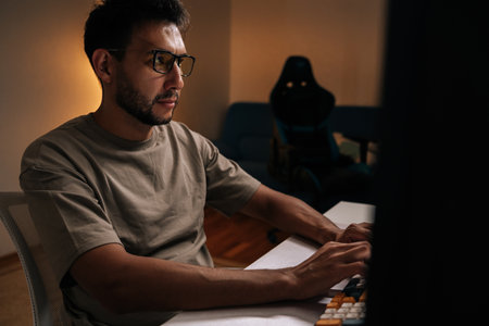 Focused programmer freelancer working on desktop computer late at night, illuminated by soft glow of...