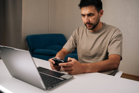 Portrait of young man concentrating playing, controlling computer game using wireless gamepad controller sitting at desk with laptop, enjoying leisure time and digital entertainment at home.の写真素材
