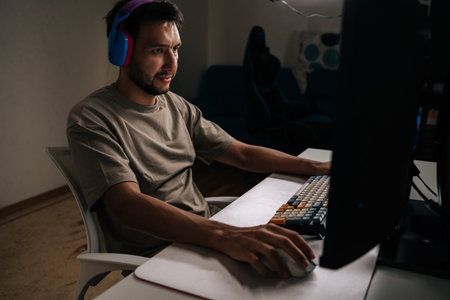 Focused young man wearing headphones and casual t-shirt, concentrating playing video game on desktop computer, enjoying immersive entertainment during leisure time at home in dark room.の写真素材