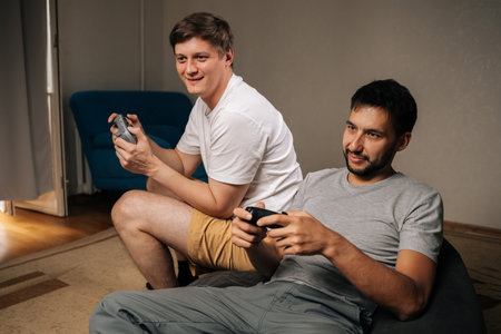 Cheerful two young men relaxing on couch, smiling and playing console video games together in cozy living room, sharing fun, competition and friendly bonding moments.の写真素材