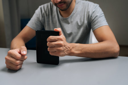 Close-up cropped shot of bearded student male enjoying digital literature, focusing on screen of ebook reader, learning and accessing information through modern technology sitting at home desk.の写真素材