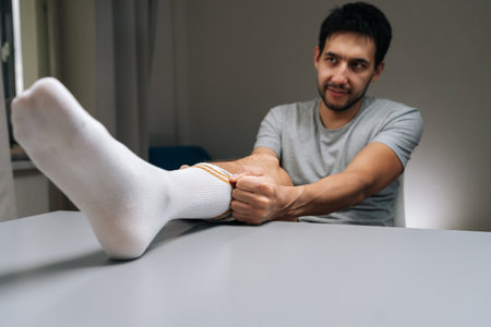 Young man putting on white sock with brown stripes sitting at table at home, expressing funny, strange, quirky feeling during daily routine.の写真素材