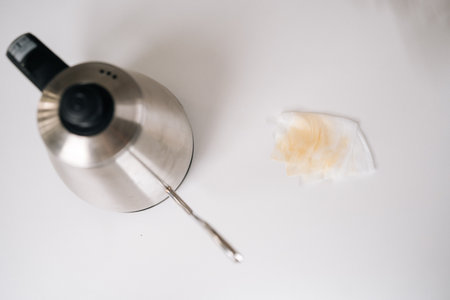High-angle view of electric kettle pouring steaming water into drip coffee maker, coffee-stained napkin nearby highlighting potential kitchen mishap on clean white surface, close-up, nobody.の写真素材