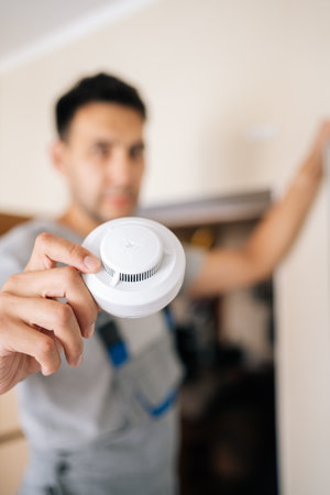 Selective focus of skilled electrician holding smoke detector while installing in residential building, prioritizing fire safety and ensuring protection for all occupants within home, looking cameraの写真素材