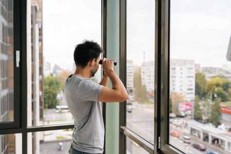 Rear view of curious young man holding monocular, looking closely out of window at urban street, engaging in observation while monitoring surroundings from apartment or home.の写真素材