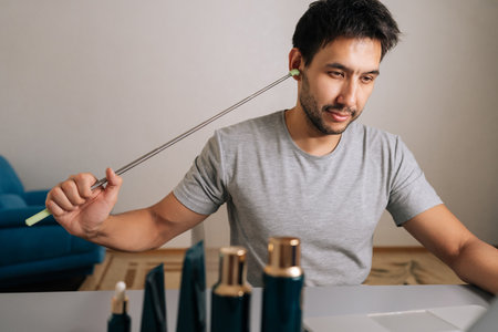Portrait of millennial man scratching ear with back scratcher working from home, browsing online for...