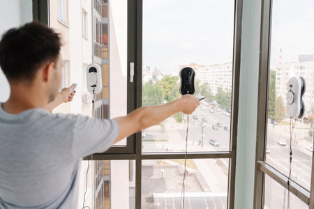 Back view of man controlling cleaning robots on skyscraper window using remote control, showcasing innovative technology for high-rise window cleaning with cityscape view. Concept of cleaning houseの写真素材