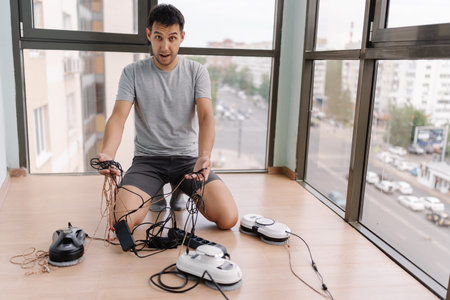Portrait of frustrated young man crouching among tangled window cleaning robot cables,...