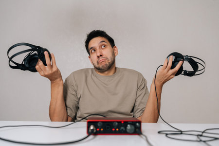 Portrait of pensive man holding two of studio headphones, looking confused and making difficult choice while sitting at desk with audio interface, representing decision-making in audio production.の写真素材