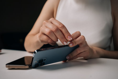 Woman sitting at white table, using sim ejector tool to open sim card tray of mobile phone, engaged in process of managing device connectivity. Concept of modern technologyの写真素材