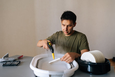 Portrait of homemaker male using steam cleaner to clean air humidifier filter in basin at home, ensuring regular maintenance for maintaining healthy indoor air quality. Concept of domestic work.の写真素材