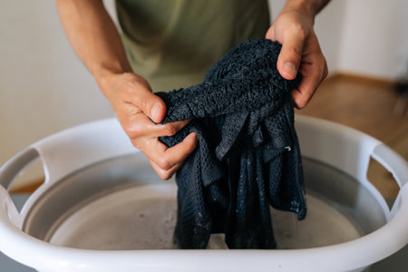 Handwashing dark clothing in plastic basin, wringing wet garment with bare hands over gray surface, showcasing manual laundry technique and water conservation. Concept of hygiene and cleanliness.の写真素材