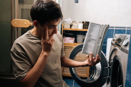 Pensive man examining dirty lint filter removed from tumble dryer, appearing concerned about large amount of lint and need for appliance cleaning and proper home maintenance.の写真素材