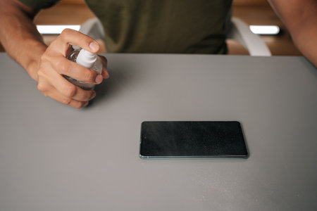 Close-up of male hands spraying sanitizer onto mobile phone, highlighting importance of hygiene and disinfection in everyday life on gray table surface. Concept of mobile phone maintenance and serviceの写真素材