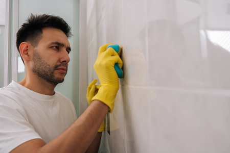 Focused bearded man cleaning bathroom tiles with blue sponge while wearing yellow rubber gloves, focusing on scrubbing white tiled wall for maintaining hygiene and completing household chores.の写真素材