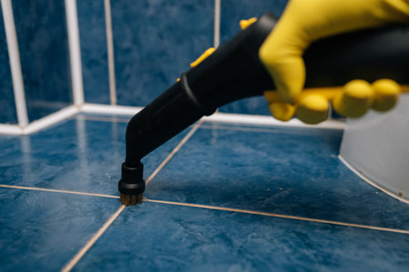 Close-up of person in yellow protective gloves operating powerful steam cleaner with brush attachment, deep cleaning blue ceramic bathroom tiles and grout lines for enhanced hygiene and sanitation.の写真素材