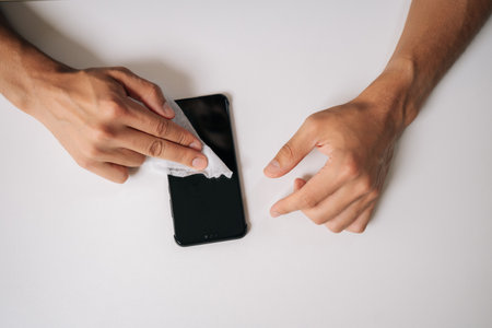 Closeup high-angle view of man carefully wiping smartphone screen with disinfectant wipe, ensuring...