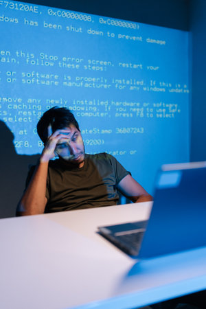 Vertical portrait of confused young male programmer facing challenging coding problem, feeling frustrated while working on laptop with lines of blue code projecting on wall behind him.の写真素材
