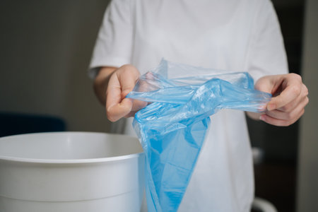Hands unfolding and positioning fresh blue plastic bag into empty white trash can, signifying start...