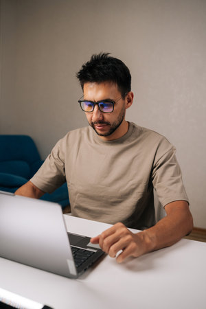 Bearded young businessman wearing glasses concentrating on work on laptop, typing on keyboard in...