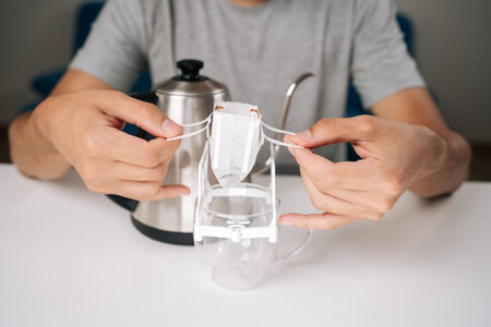 Close-up cropped shot of unrecognizable man placing drip coffee bag onto glass cup, showcasing process of brewing freshly ground coffee with kettle in background. Concept of enjoying morning ritual.の写真素材