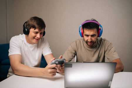 Happy excited young men gathered in living room, laughing and focused on laptop and controllers...