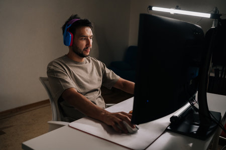 Gamer male wearing colorful headphones engrossed in playing challenging video game on personal...