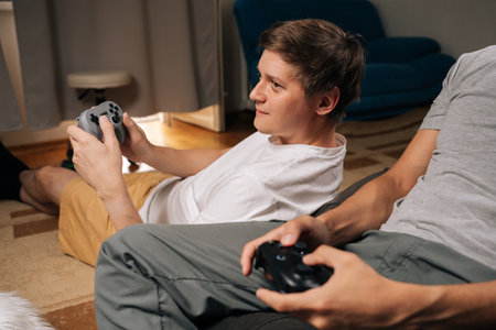 Two young adult male friends sitting comfortably at home, deeply engaged in playing video game...